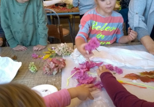 Dzieci siedzą przy stole. Dwie dziewczynki smarują kartkę klejem. Pozostałe dzieci przeklejają kwiaty do zarysu postaci anioła.
